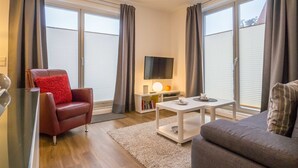 A modern living room with a leather armchair, a grey sofa, a white coffee table, and a flat-screen TV. - App. 02 / EC - Apartment No. 02 in the sunbath (Sassnitz)