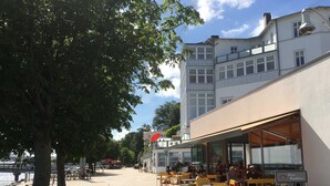 An outdoor café with yellow chairs and tables, a view of the water, and a building with large windows in the background. - App. 02 / EC - Apartment No. 02 in the sunbath (Sassnitz)