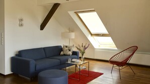 Un salon moderne avec un canapé bleu, un fauteuil rouge, une table basse en verre et une lucarne.