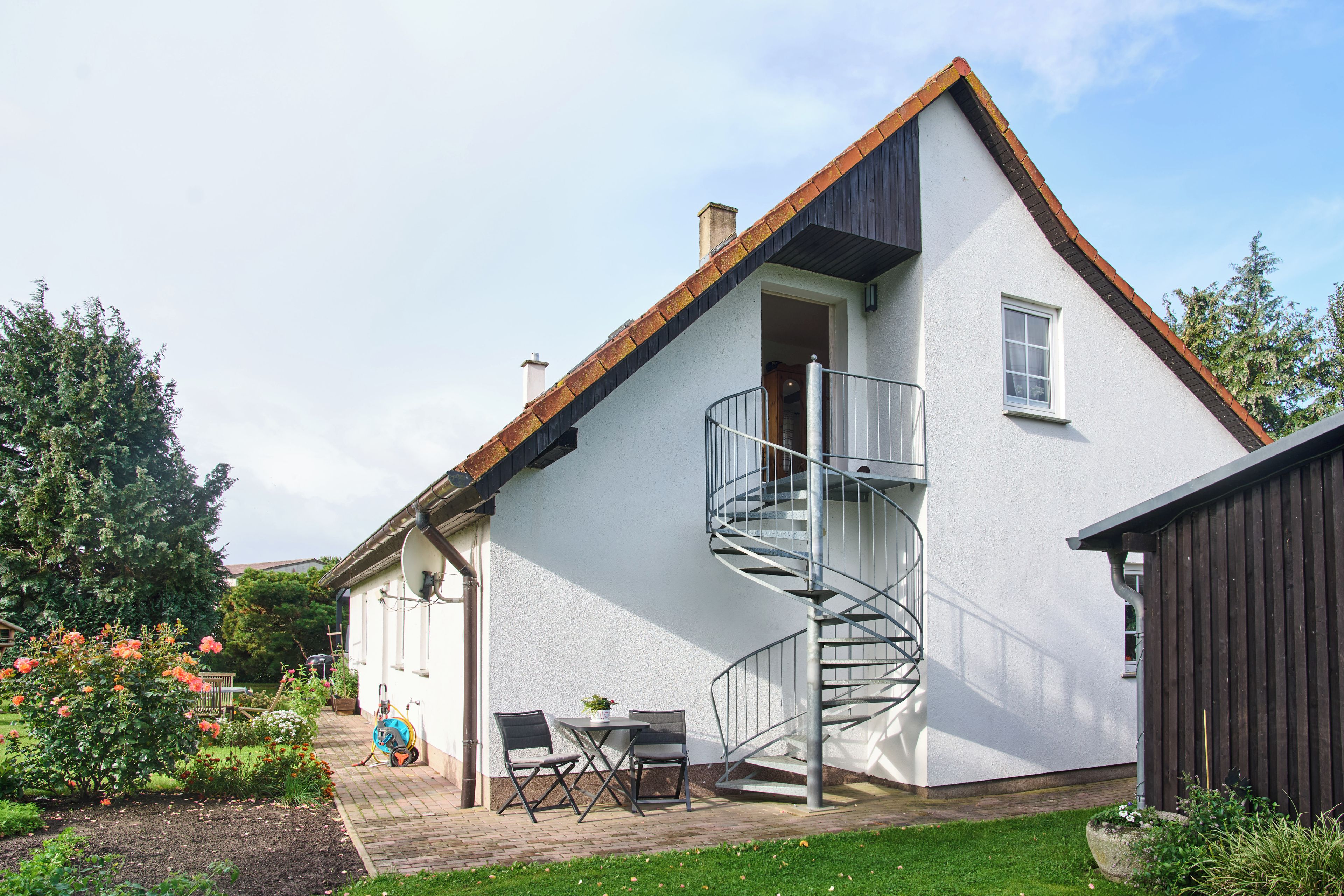 A two-story house with a spiral staircase, a patio with outdoor furniture, and a garden with flowering plants.