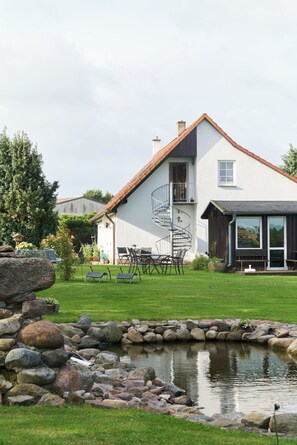 A two-story house with a white exterior, a visible balcony, and a small pond in the foreground. - Peace and relaxation in a restful natural setting (Rappin OT Helle)