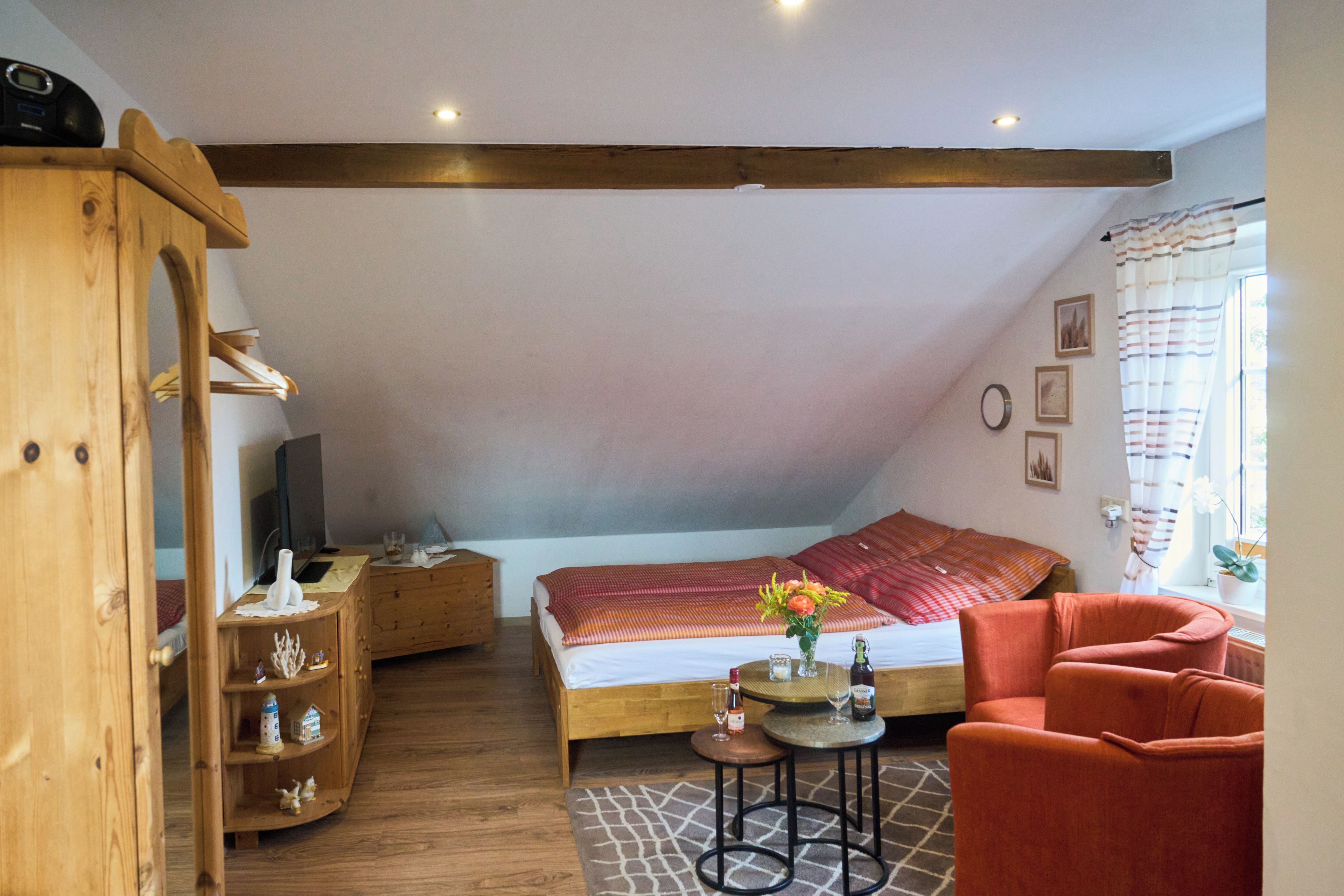 Une chambre mansardée chaleureuse, avec un plafond en pente, une armoire en bois, un lit recouvert d’une couverture à rayures rouges et oranges, une petite table ronde ornée d’un vase et de bouteilles, et un fauteuil rouge.