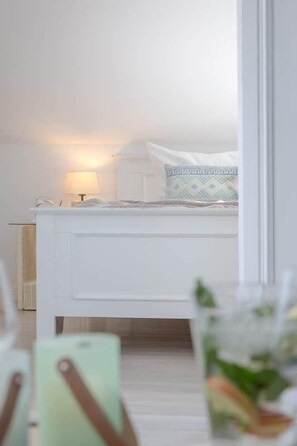 A white bed with a patterned pillow, a bedside lamp, and a clear glass with a plant.