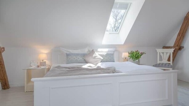 A bedroom with a white bed, a skylight, a bedside table with a lamp, and a small plant. - Vacation apartment Watthof (Emmelsbüll-Horsbüll)