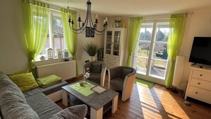 Un salon comprenant un canapé, un fauteuil, une table basse et un téléviseur. On y trouve des rideaux verts, un lustre et un meuble avec des portes en verre.
