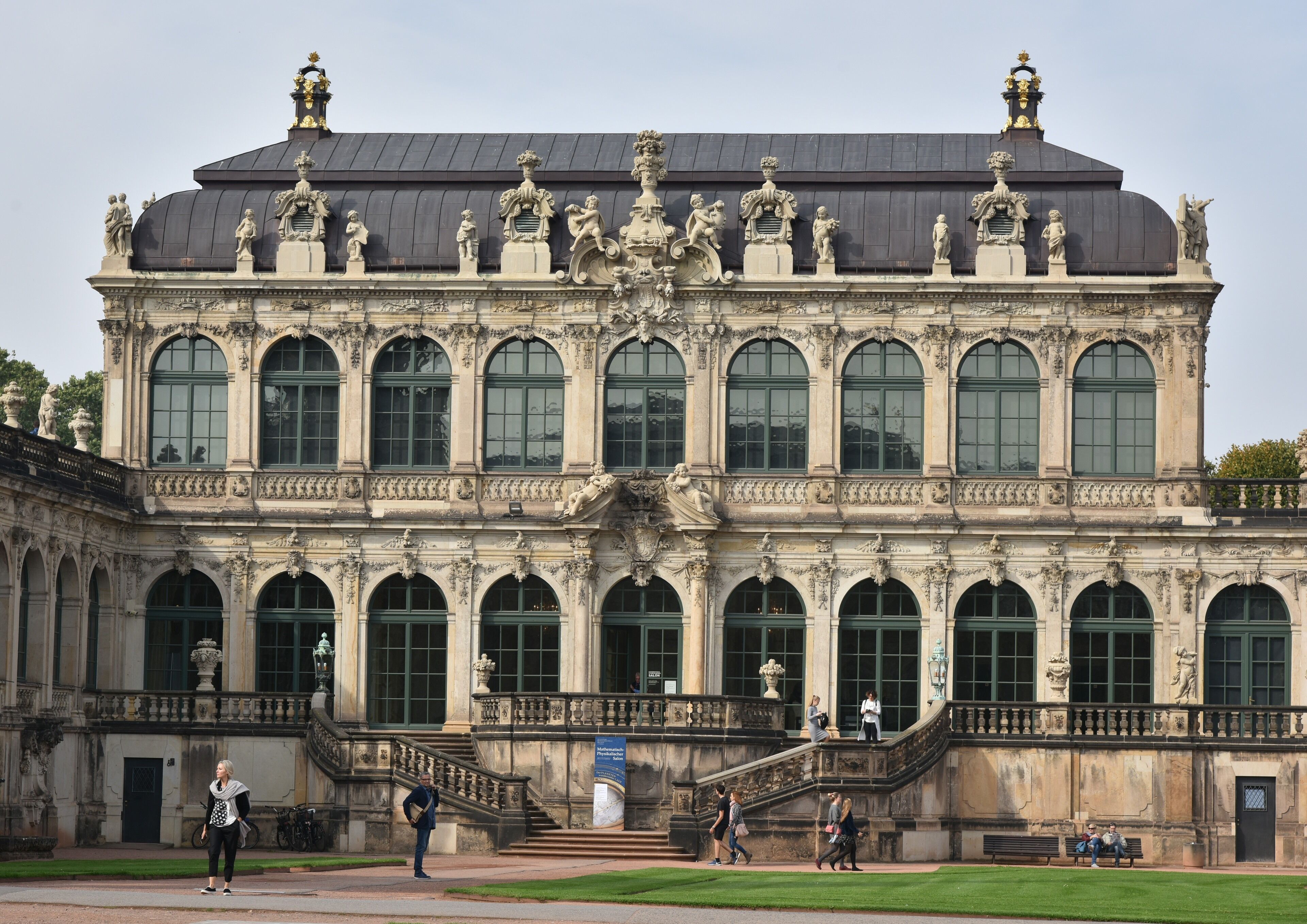 Ein prachtvolles Gebäude mit klassischer Architektur, das eine zentrale Kuppel, mehrere Rundbogenfenster und Statuen auf dem Dach zeigt.