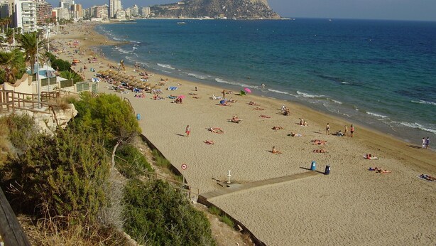 Une plage avec une formation rocheuse imposante en arrière-plan, un rivage sablonneux et des bâtiments le long de la côte.