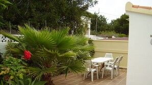 Une terrasse avec une table et des chaises blanches, entourée de plantes vertes et d’une fleur rouge.