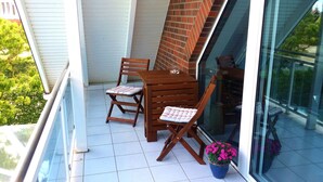 Ein Balkon mit Holztisch, zwei Stühlen, einem Blumentopf und einer Glastür, die in einen Garten führt.