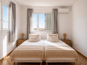 A double bed with a patterned bedspread, two bedside tables with lamps, a window with curtains, and an air conditioning unit. - Private Family Villa with Pool & Garden, 3km from Beach, Relaxing Location (Rhodos)