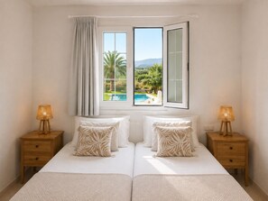 A double bed with a view of a swimming pool and mountains through an open window.