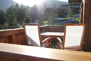 Eine Holzterrasse mit zwei Stühlen und einem Tisch, mit Blick auf eine gebirgige Landschaft mit Bäumen und Gebäuden.