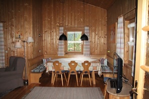 Ein Holzblockhaus-Innenraum mit Esstisch, drei Stühlen, einer Couch, einem Fernseher und einem Fenster mit Vorhängen.