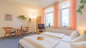 Une chambre d’hôtel avec deux lits, une télévision, une table à manger avec des chaises, une plante et une fenêtre avec des rideaux.