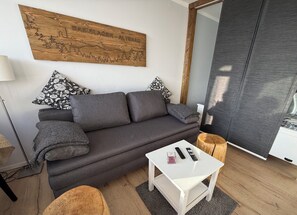 Una sala de estar moderna con un sofá gris, dos almohadas estampadas, una mesa de centro blanca con controles remotos y una obra de arte de madera en la pared.