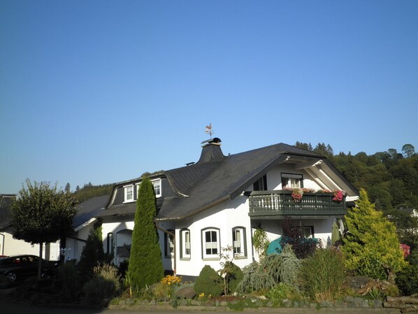 A two-story house with a steeply pitched roof, a balcony with hanging flower baskets, and a garden with various plants and flowers. - ☀️☀️☀️ cozy apartment with additional room to expand (Willingen)
