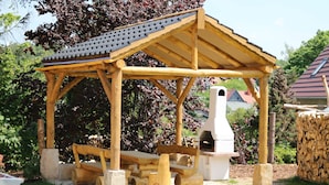 Ein hölzernes Gartenhaus mit geneigtem Dach, einer Steinsitzbank und einem Holzofen.