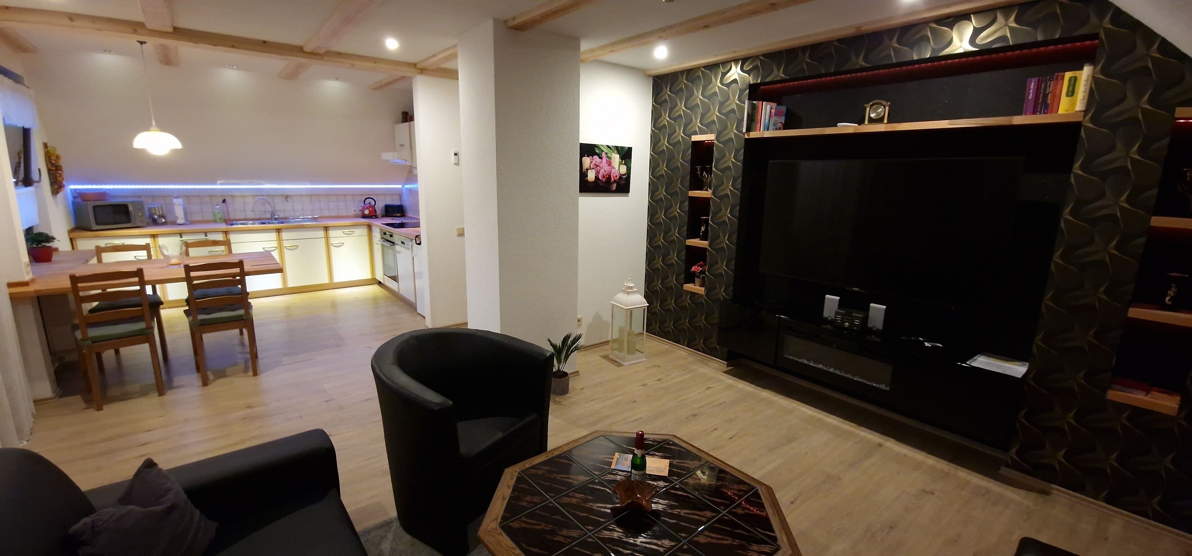 Ein modernes Wohnzimmer mit einer Couch, einem Couchtisch und einem Fernseher. Im Hintergrund ist eine Küche mit einem Esstisch und Stühlen zu sehen.