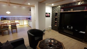 Ein modernes Wohnzimmer mit einer Couch, einem Couchtisch und einem Fernseher. Im Hintergrund ist eine Küche mit einem Esstisch und Stühlen zu sehen.