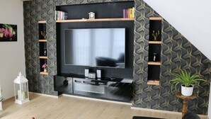 Ein modernes Wohnzimmer mit einem großen Flachbildfernseher, Holzregalen und einer Topfpflanze.