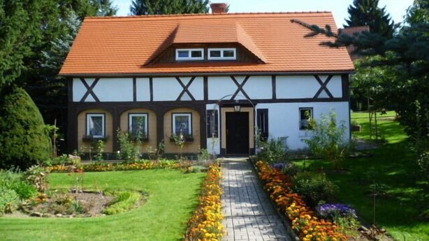 Ein traditionelles Fachwerkhäuschen mit rotem Ziegeldach, einer Haustür und einem Garten mit gepflastertem Weg und Blumenbeeten.