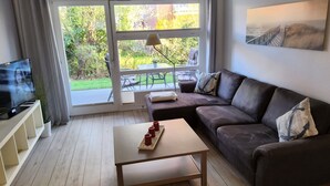 A living room with a sofa, a coffee table, a TV, and a view of a garden through a sliding door. - Renovated 2-room apartment, bright and friendly, dogs are welcome (Schönberg)