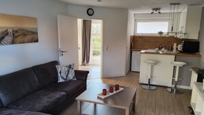 A compact living space with a sofa, a coffee table, a kitchen area with a bar stool, and a door leading to an outdoor area. - Renovated 2-room apartment, bright and friendly, dogs are welcome (Schönberg)