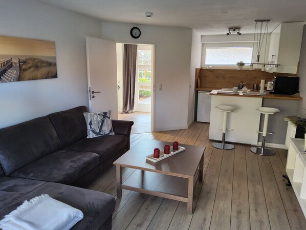 Un espacio habitable compacto con un sofá, una mesa de centro, una zona de cocina con un taburete alto y una puerta que da a un área exterior.