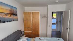 A bedroom with a bed, a painting of a beach scene, a wooden wardrobe, and a hallway leading to a door. - Renovated 2-room apartment, bright and friendly, dogs are welcome (Schönberg)