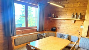 Interior de una cabaña de madera con una gran mesa de madera, bancos acolchados y una estantería con botellas.