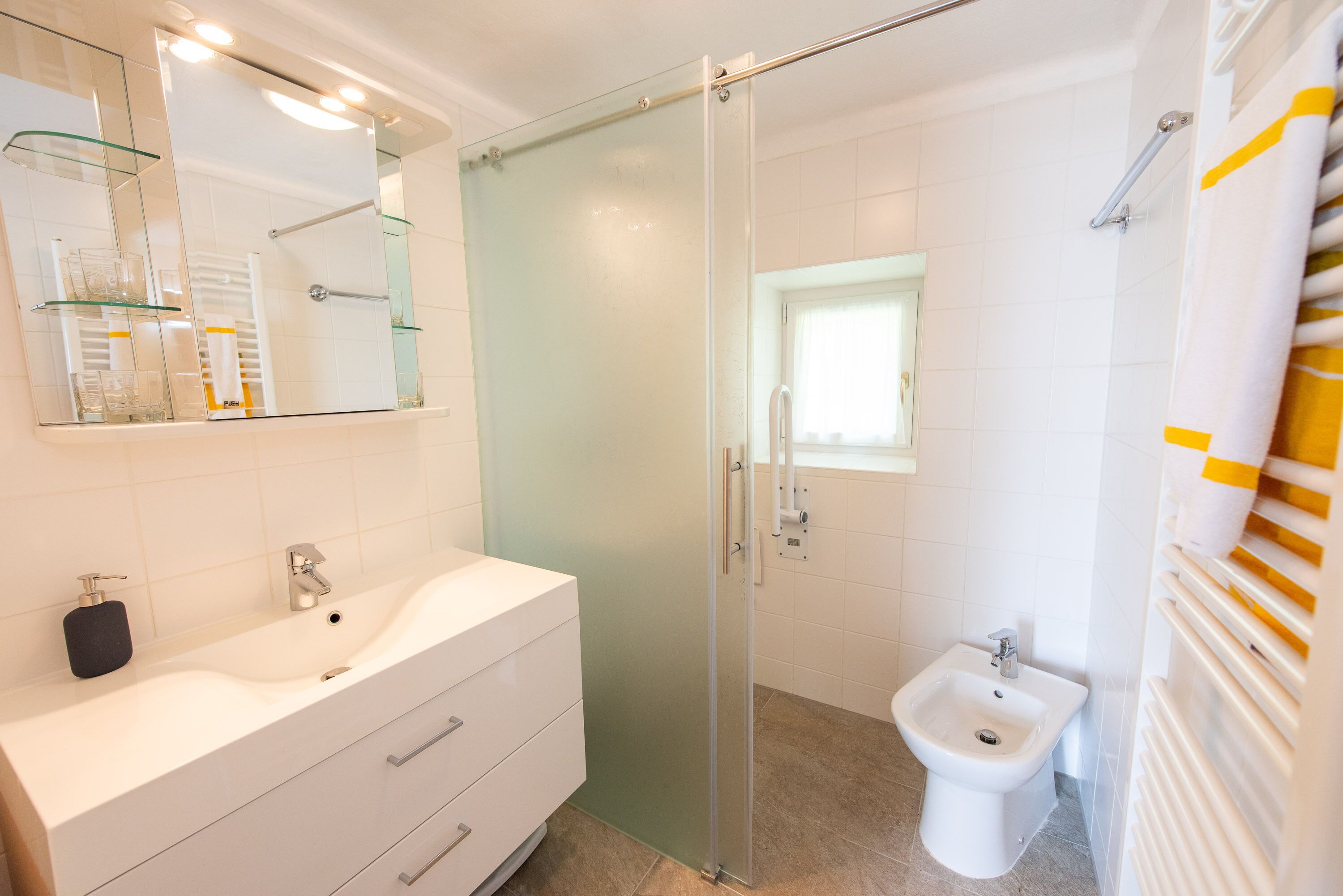 A modern bathroom with a glass-enclosed shower, a white sink with a soap dispenser, and a white pedestal bidet.