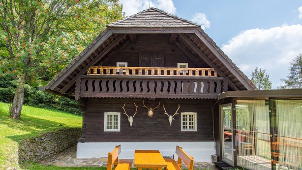 A wooden cabin with a balcony, a dining table with benches, and a stone wall.