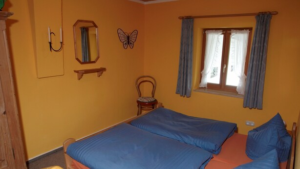 Un dormitorio con dos camas, una silla de madera, una mesita, un espejo, un cuadro de mariposas y una ventana con cortinas.