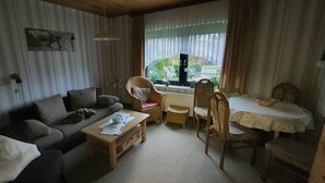 A cozy living room with a sofa, a wooden coffee table, a dining table with chairs, and a window with curtains. - Small and cozy holiday home. Quiet Location (Bunde)