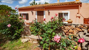 Une maison de campagne avec un mur en pierre, un toit de tuiles et un jardin où poussent des rosiers roses.