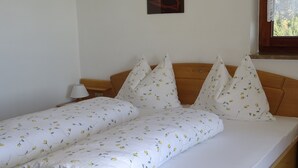 Cama com colcha florida e várias almofadas, cabeceira de madeira, mesa de cabeceira com abajur e um quadro na parede.