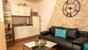 Un espace de vie compact comprenant un coin cuisine, un canapé en cuir noir agrémenté d’un coussin bleu et une horloge murale.