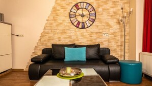 Un salon avec un canapé en cuir noir, une horloge murale ronde, une table basse et un pouf bleu.