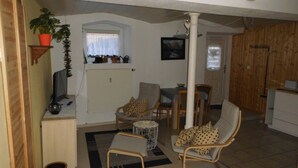 Ein gemütliches Wohnzimmer mit einer Couch, Sesseln und einem kleinen Couchtisch. Ein Fenster mit Vorhängen und eine Heizung sind sichtbar.