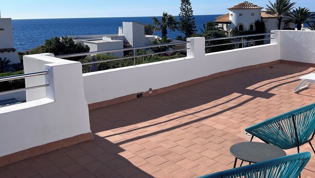 A rooftop terrace with a view of the sea, featuring a white building, palm trees, and two blue chairs.