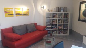A red sofa with grey cushions, a glass coffee table with a vase, and a bookshelf filled with books and decorative items. - High-quality-modern, approx 65sqm, gr. Garden, exclusive equipment, wirkl. gr. bathroom (Lüneburg)