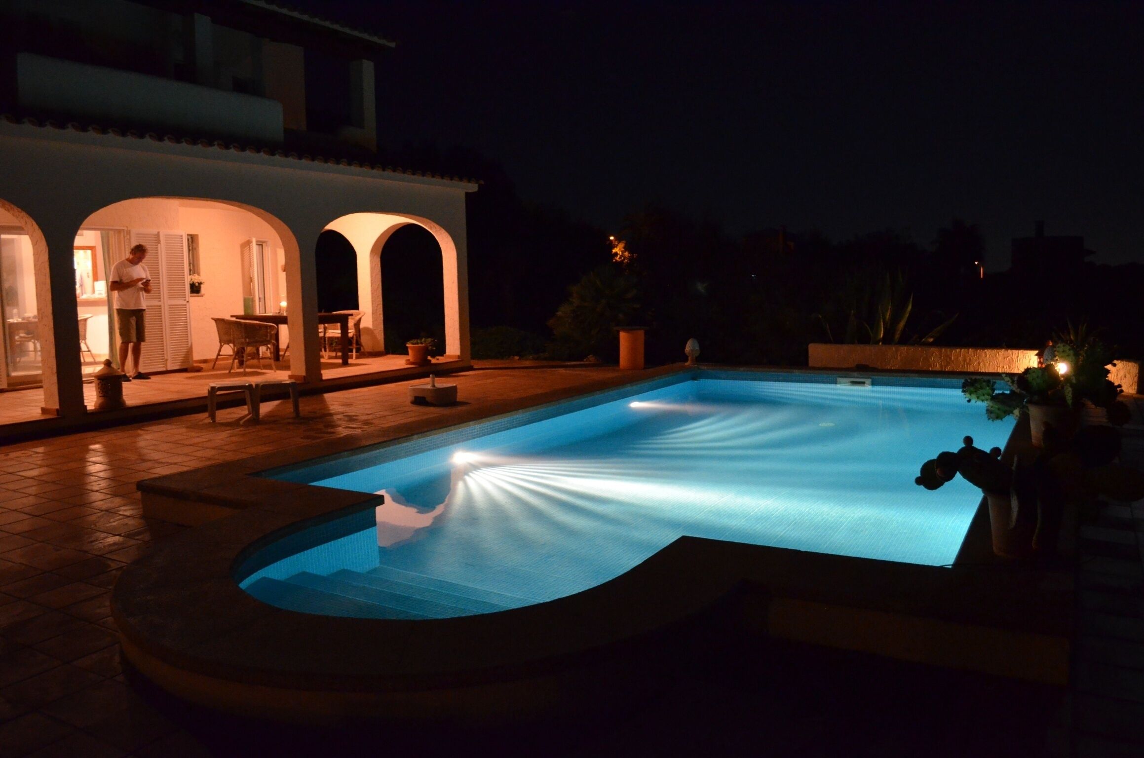 A swimming pool with a curved edge, a patio area with a table and chairs, and a building with arched doorways.