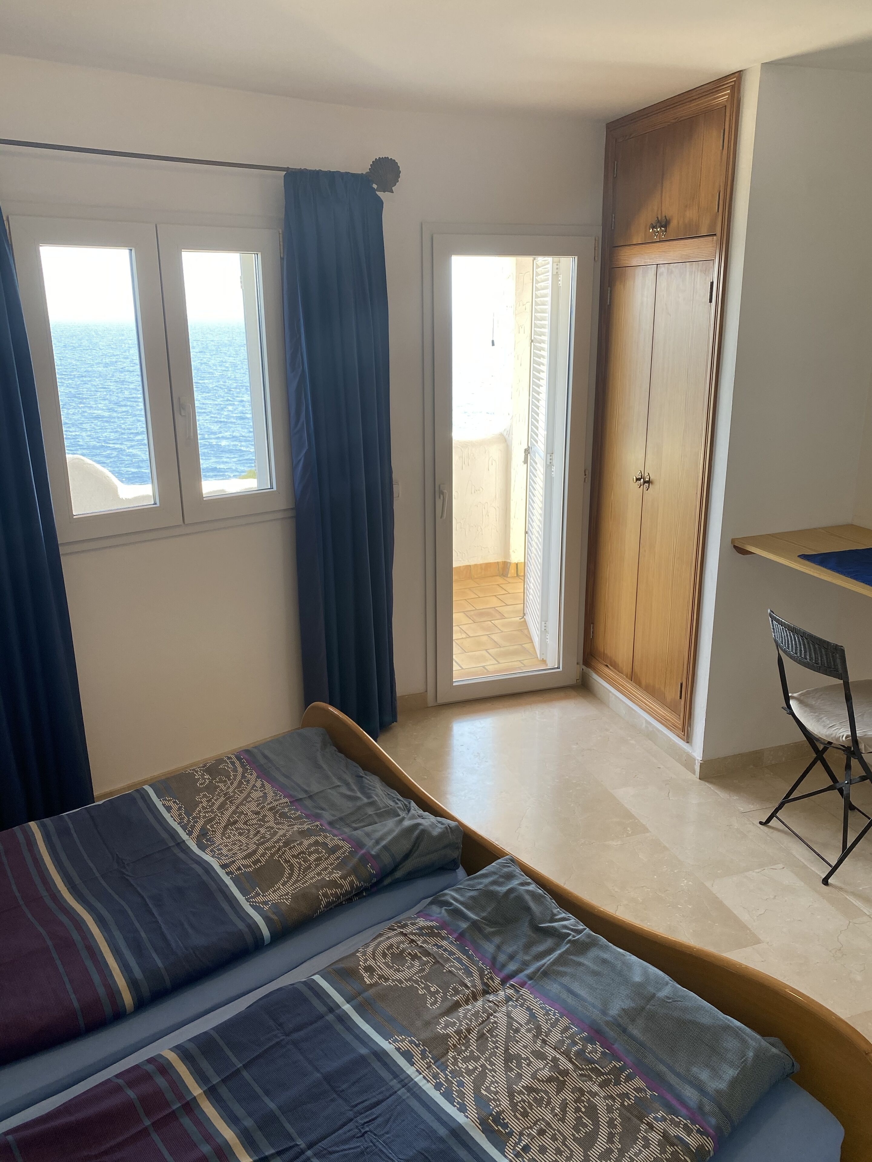 Une chambre à coucher comprenant un lit, un bureau, une chaise, une armoire et offrant une vue sur la mer par les fenêtres.