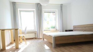 A bedroom with a wooden bed, a small table, and a chair. There are two windows with curtains, and the room has wooden flooring. - 2 room cozy Apartment - Sunny ground floor apartment with garden terrace in Forchheim (Forchheim)