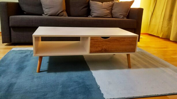 A living room with a white coffee table with a drawer, a dark grey sofa with cushions, and a blue and white rug. - 2 room cozy Apartment - Sunny ground floor apartment with garden terrace in Forchheim (Forchheim)