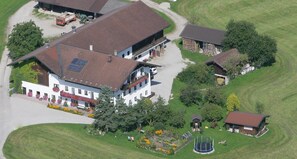 Un edificio grande con varias secciones, incluyendo un cuerpo principal con techo inclinado y una estructura más pequeña adyacente. Hay un camino pavimentado que conduce a la entrada principal y un área de juegos con un trampolín.