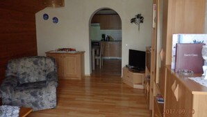 Una sala de estar con un sofá, un televisor y un aparador de madera. Se observa una cocina al fondo.