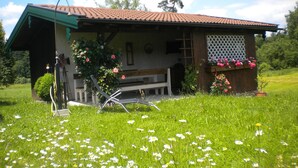 Una cabaña de madera rústica con techo inclinado, una banca afuera y un jardín con flores y césped.