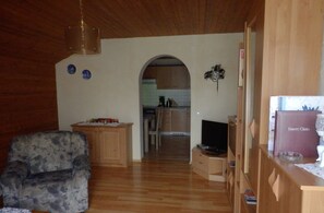 Una sala de estar con un sofá, un televisor y un aparador de madera. Se observa una cocina al fondo.