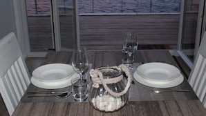 Mesa de comedor dispuesta con platos blancos, vasos y cubiertos, con un tarro de vidrio en el centro, con vista a una terraza de madera y al agua.